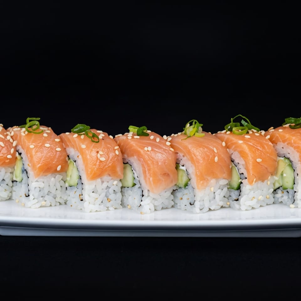 Salmon-topped sushi rolls with avocado, sesame seeds, and scallions arranged on a white rectangular plate against a black background.
