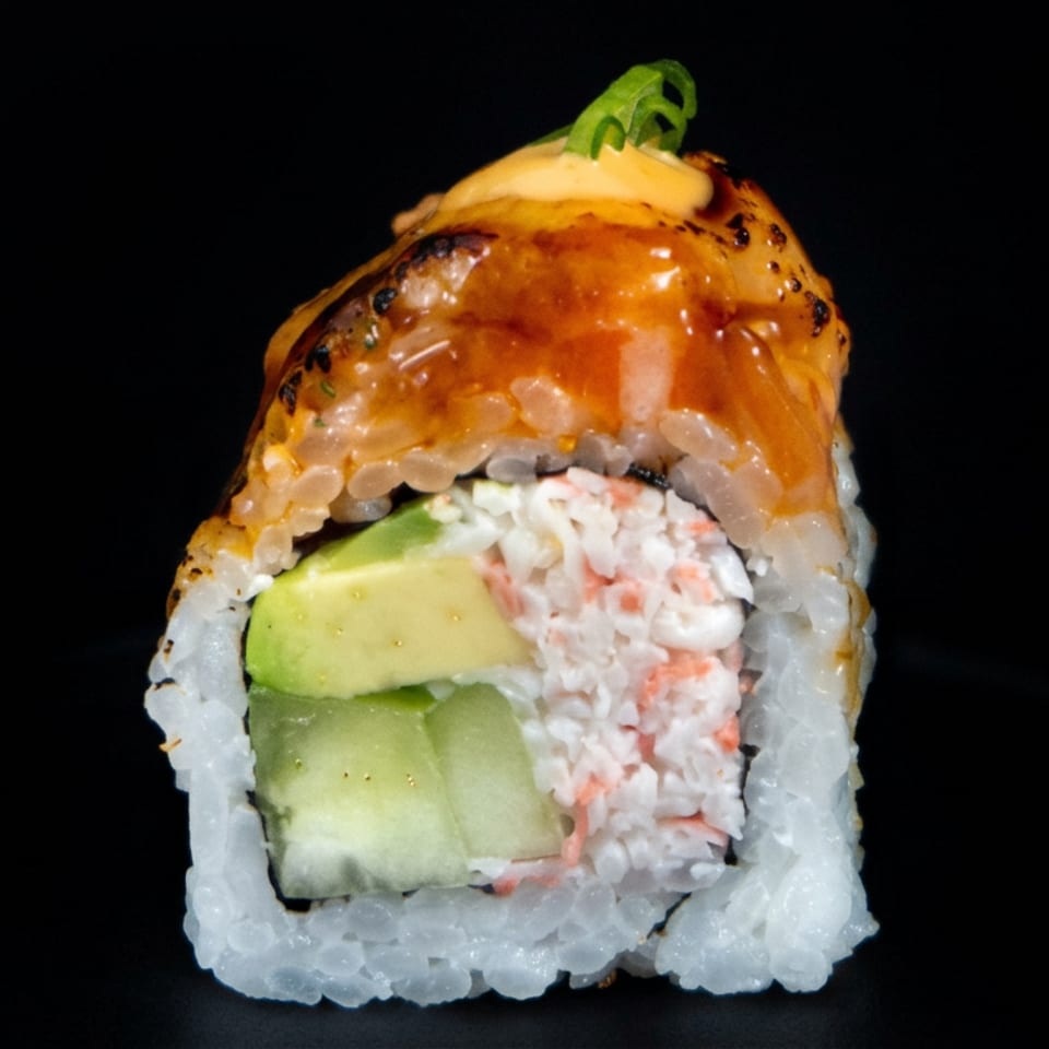 Close-up of a sushi roll filled with avocado and crab, topped with glossy orange sauce and green garnish.